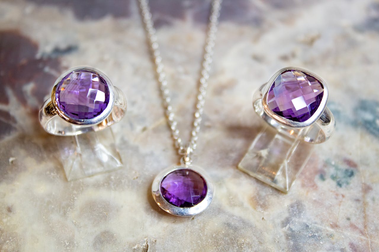 about-us Close-up of amethyst rings and necklace set on a textured surface, showcasing silver accents.